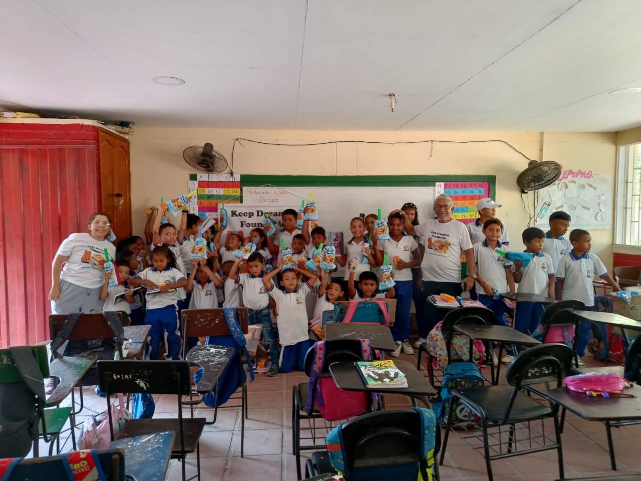 A full classroom in Colombia: children in white-and-blue uniforms raising Keep Dreaming Foundation gift bags above their heads, with two volunteers in KDF t-shirts