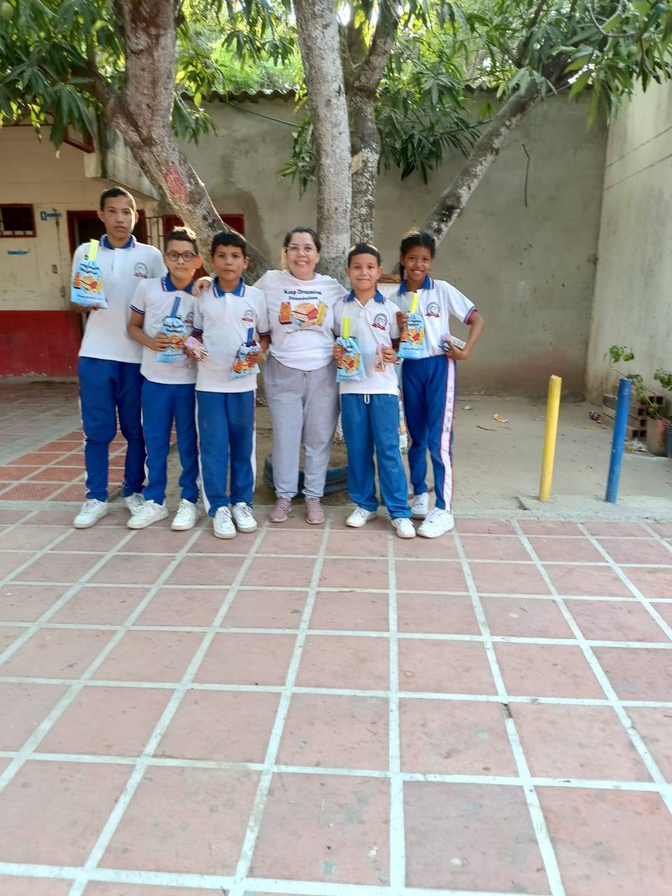 Marta in a KDF t-shirt with five schoolchildren under mango trees in the courtyard, each holding a gift bag